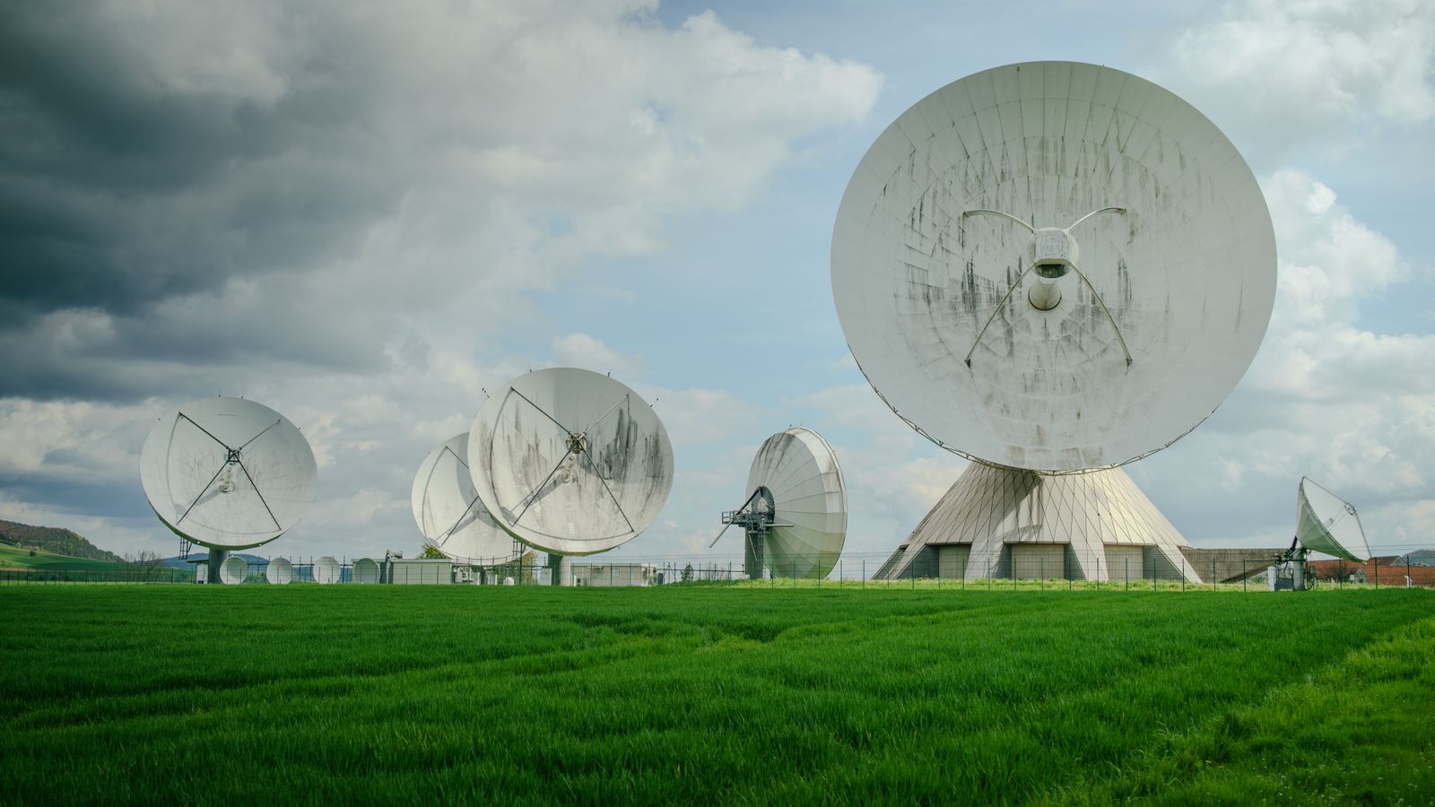 Array of large satellite dishes in a lush green field under a cloudy sky.