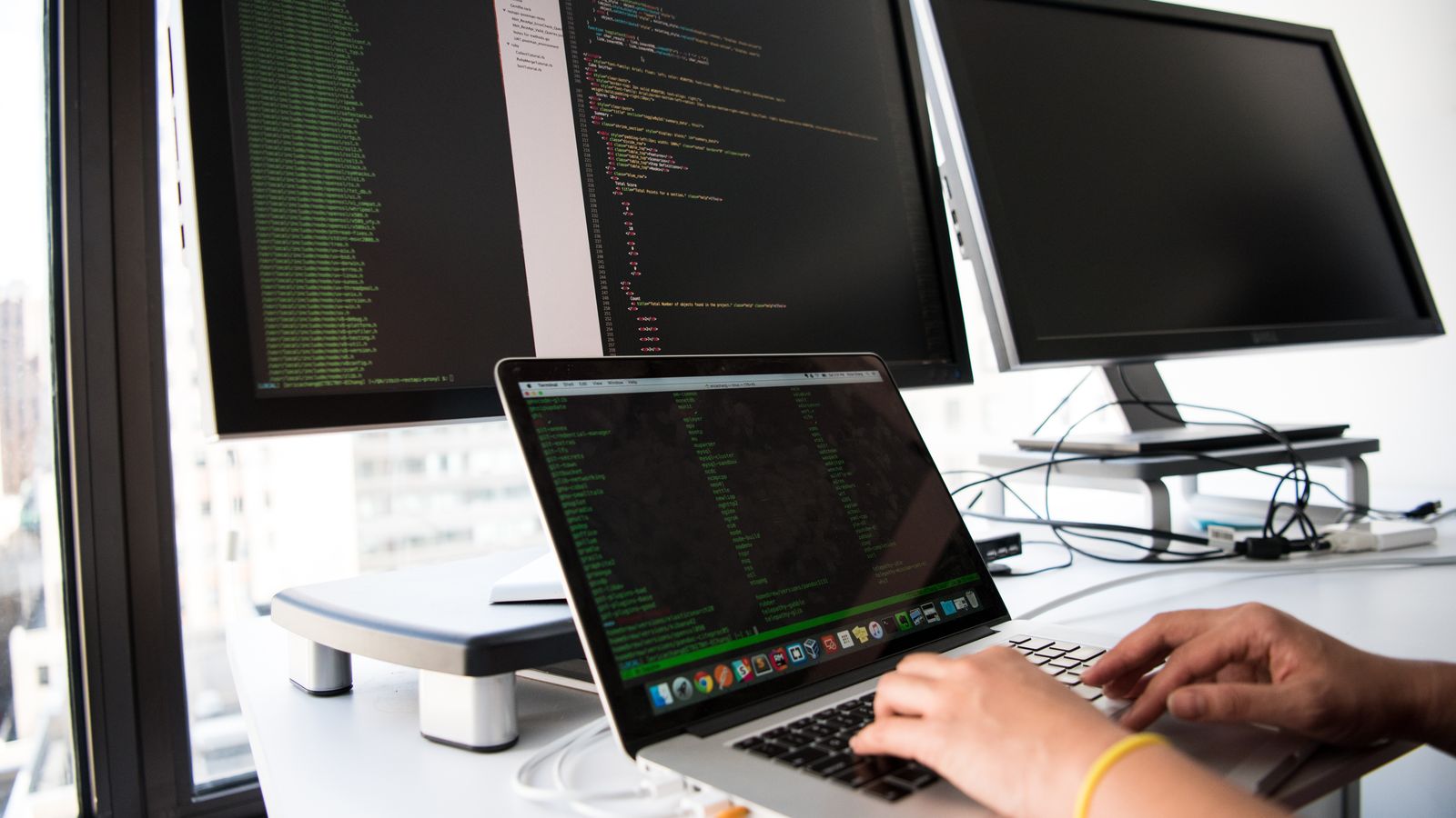 A developer writes code on a laptop in front of multiple monitors in an office setting.