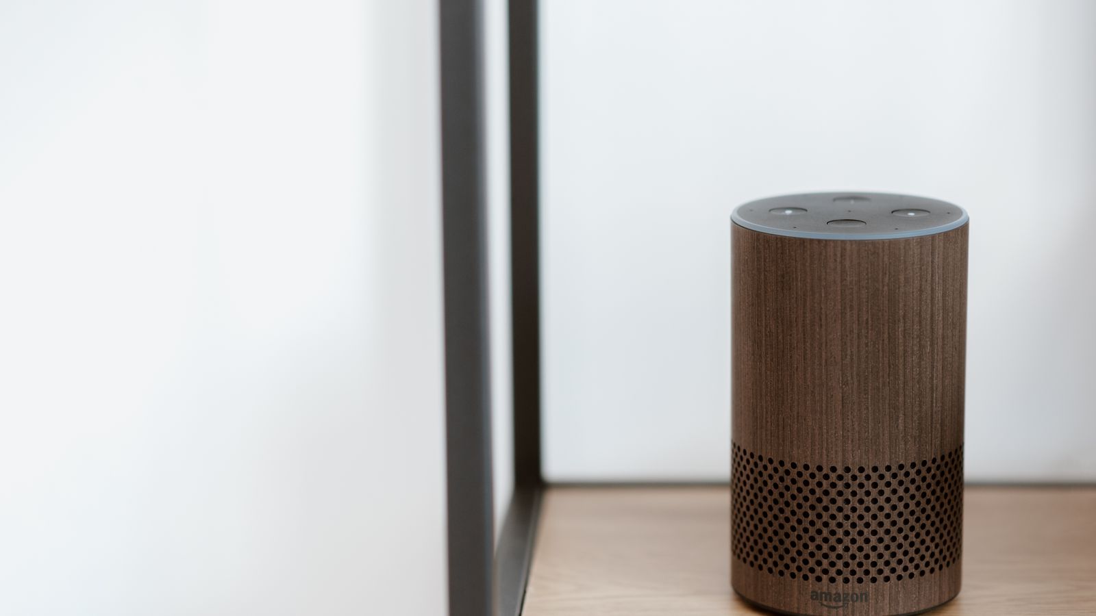 A sleek, modern smart speaker displayed on a wooden shelf in a minimalist room setting.