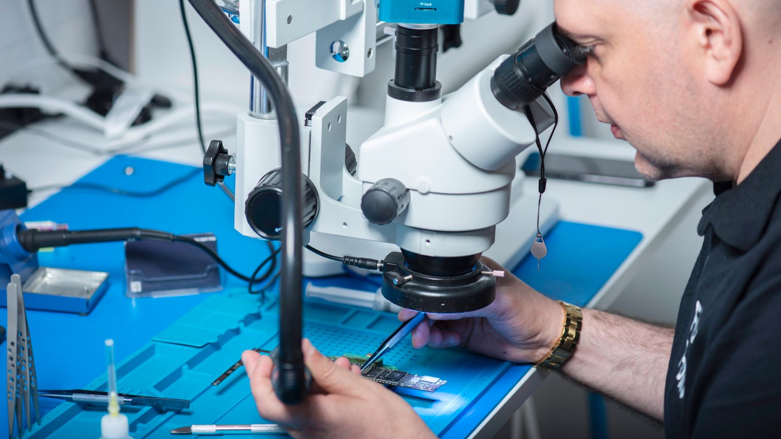 Technician examining electronic chip under a microscope in a computer lab setting.