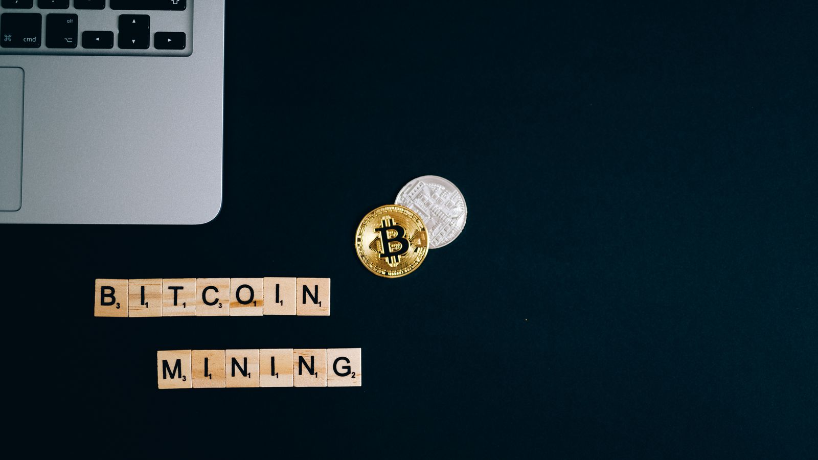 Top view of a laptop with Bitcoin coins and blocks spelling 'Bitcoin Mining' on a dark surface.