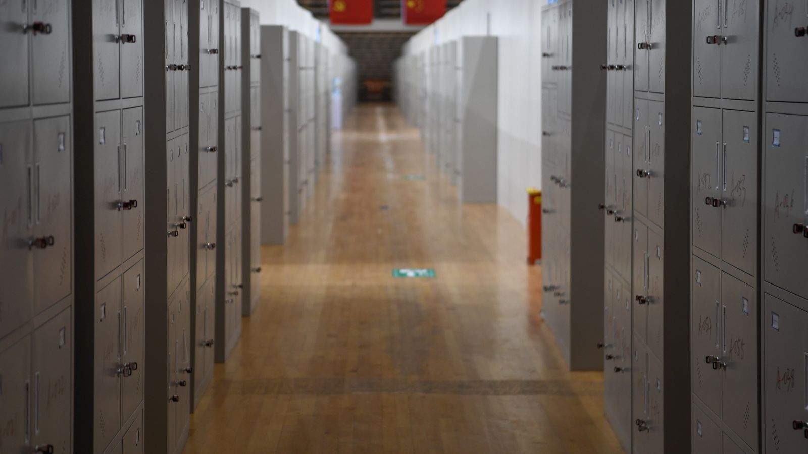 A perspective view of a long hallway featuring rows of metal lockers and wooden flooring.