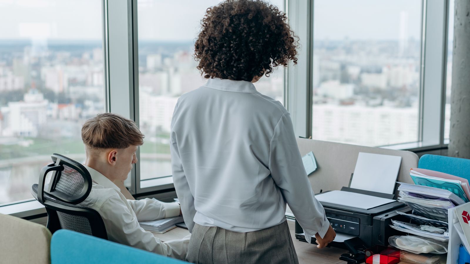 Two colleagues working in a modern office with a printer near a large window.