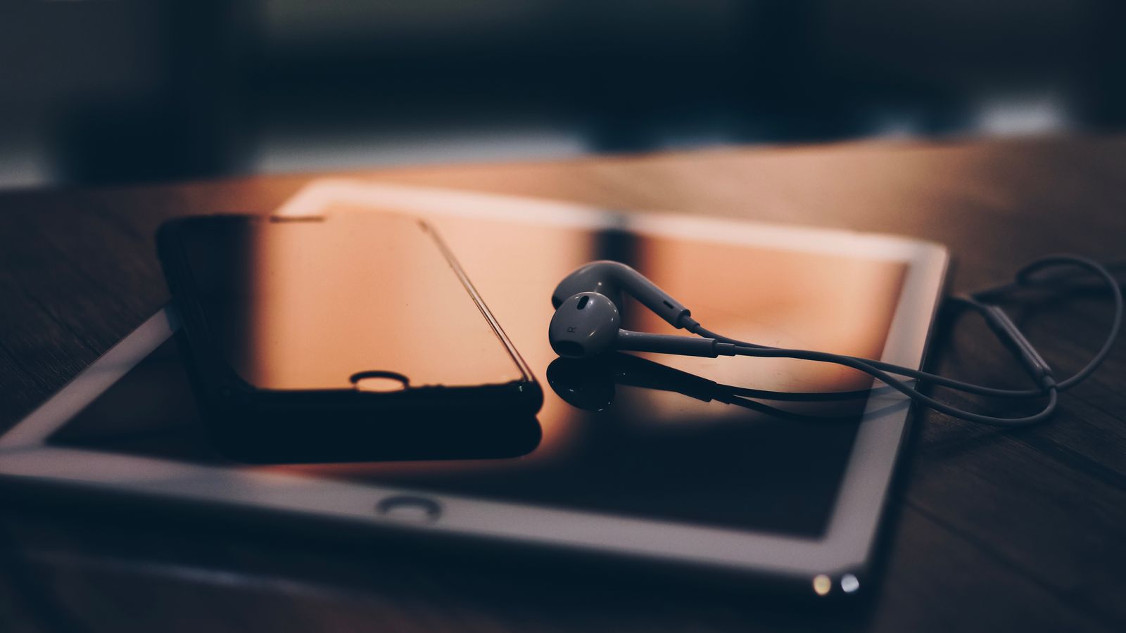 Close-up of smartphone, tablet, and earphones on a wooden table, bathed in warm lighting.