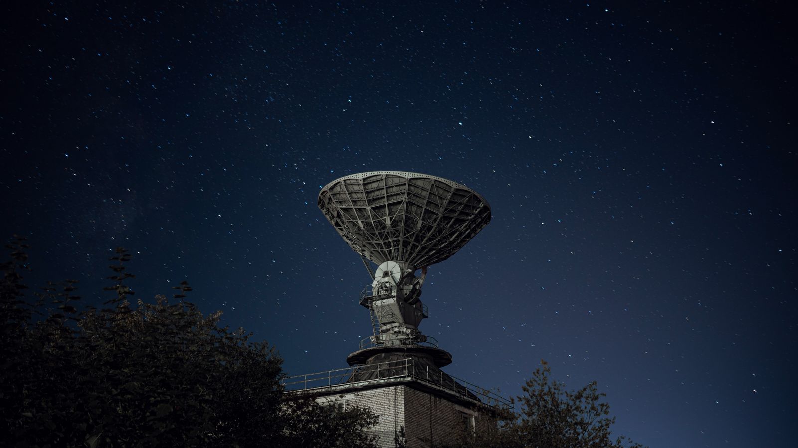 Radio telescope and green lush trees under blue sky with stars at night