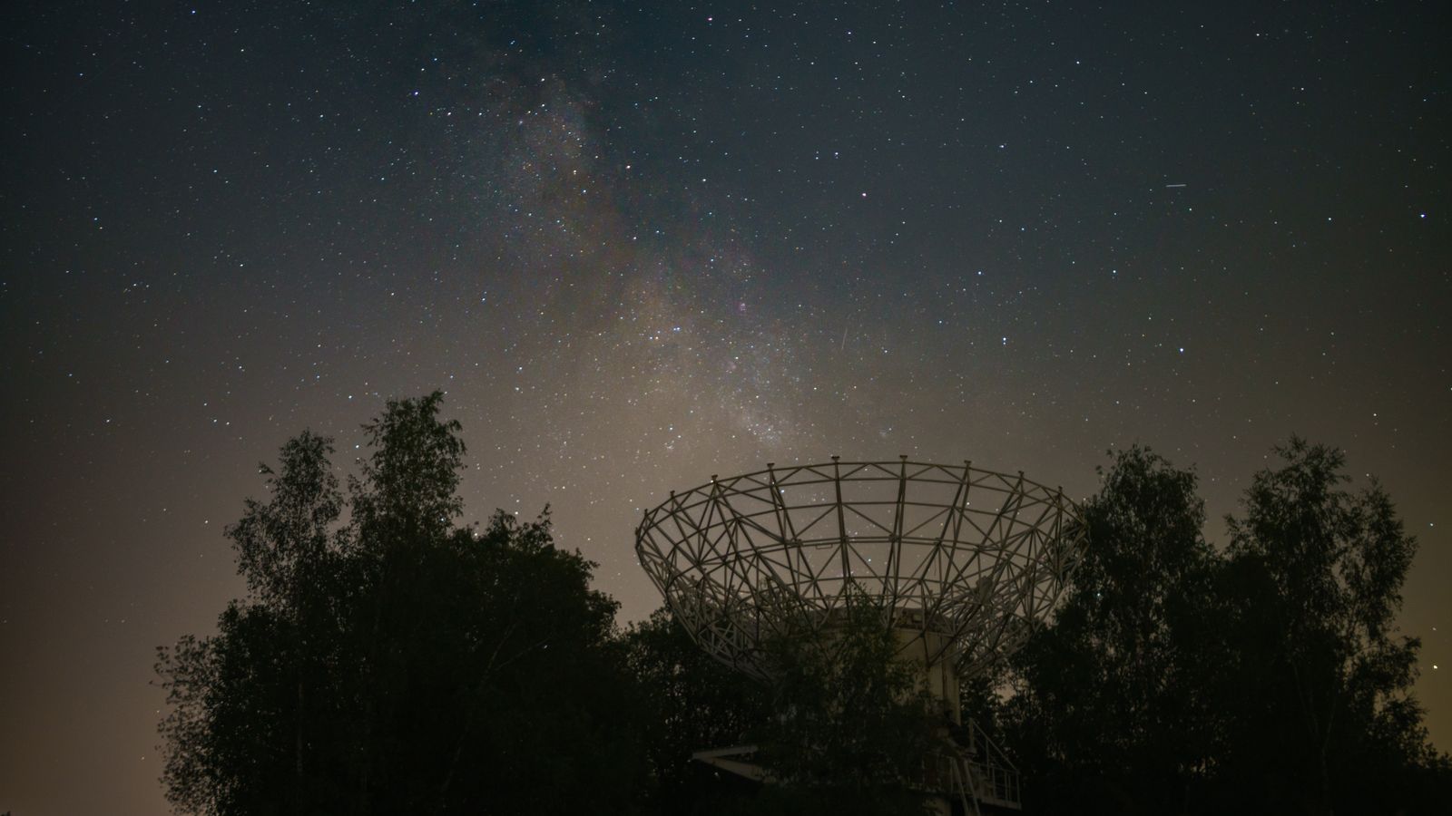 Starry night sky with radio telescope silhouette and Milky Way galaxy in view.