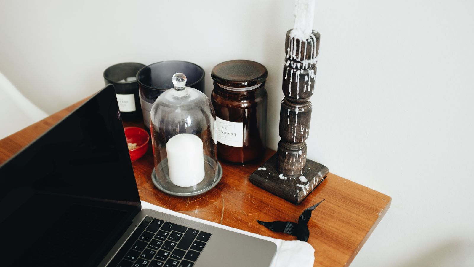 Freelance workspace with laptop and various black aromatic candles in glass jars and wooden candlestick placed on wooden table in light room of modern apartment
