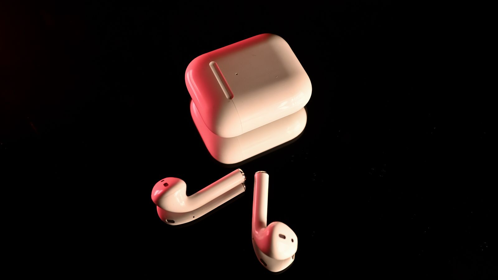 White wireless earbuds with a charging case, highlighted with red lighting on a reflective black surface.