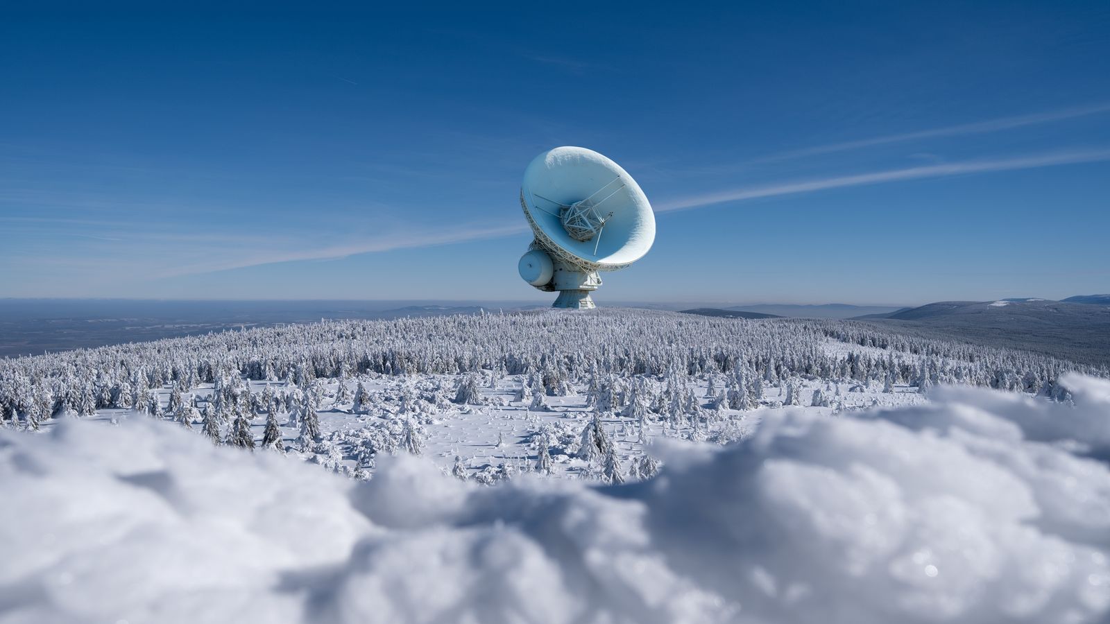 A large satellite dish observatory surrounded by snowy landscape and winter forest under a clear blue sky.