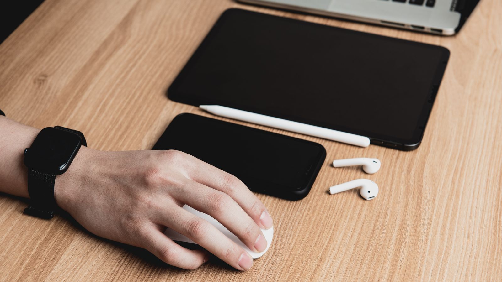 A hand using a mouse with a laptop, tablet, smartphone, and earphones on a wooden desk.