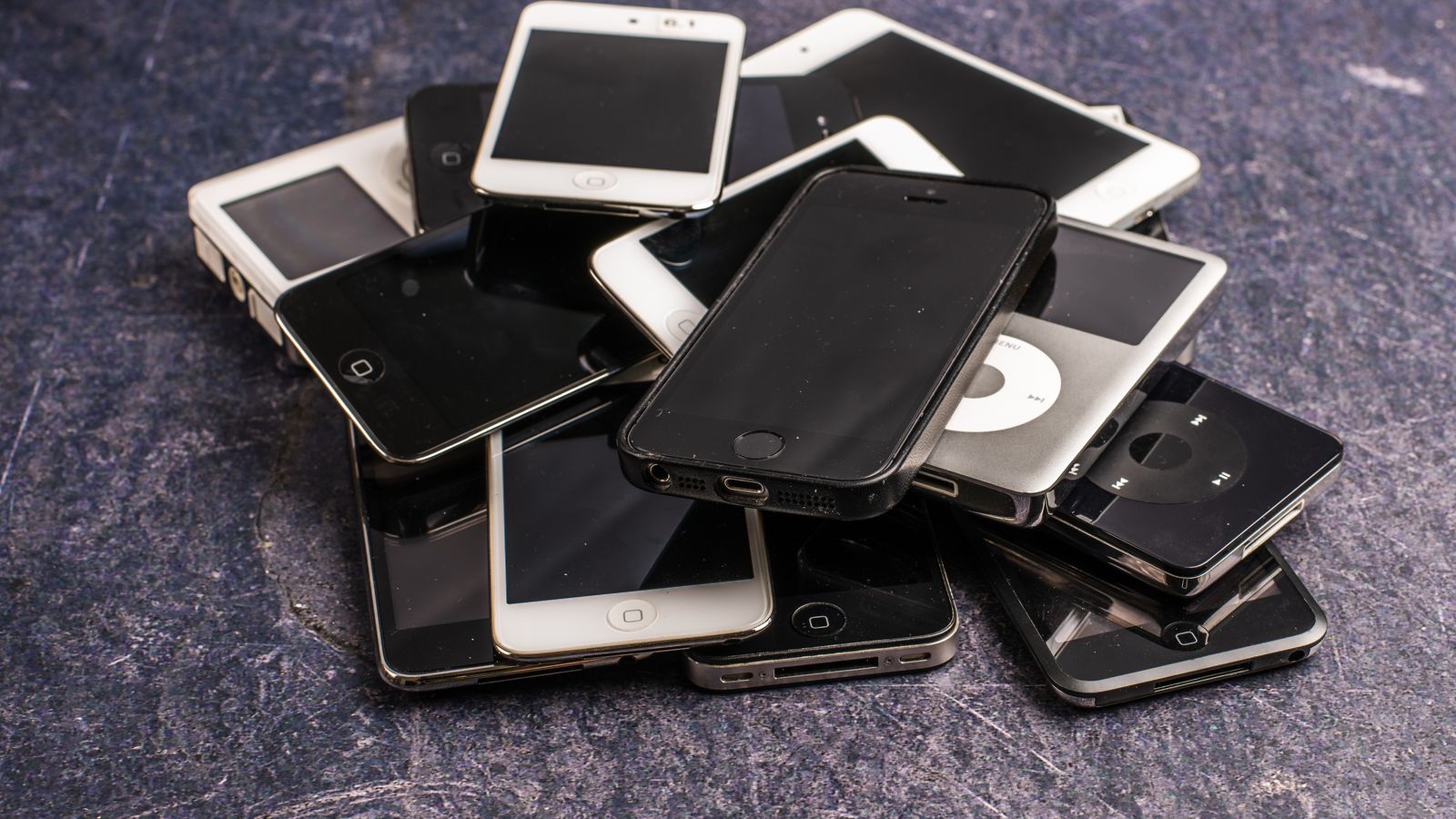 Stack of assorted aged cellphones with black screens and portable music players on gray background