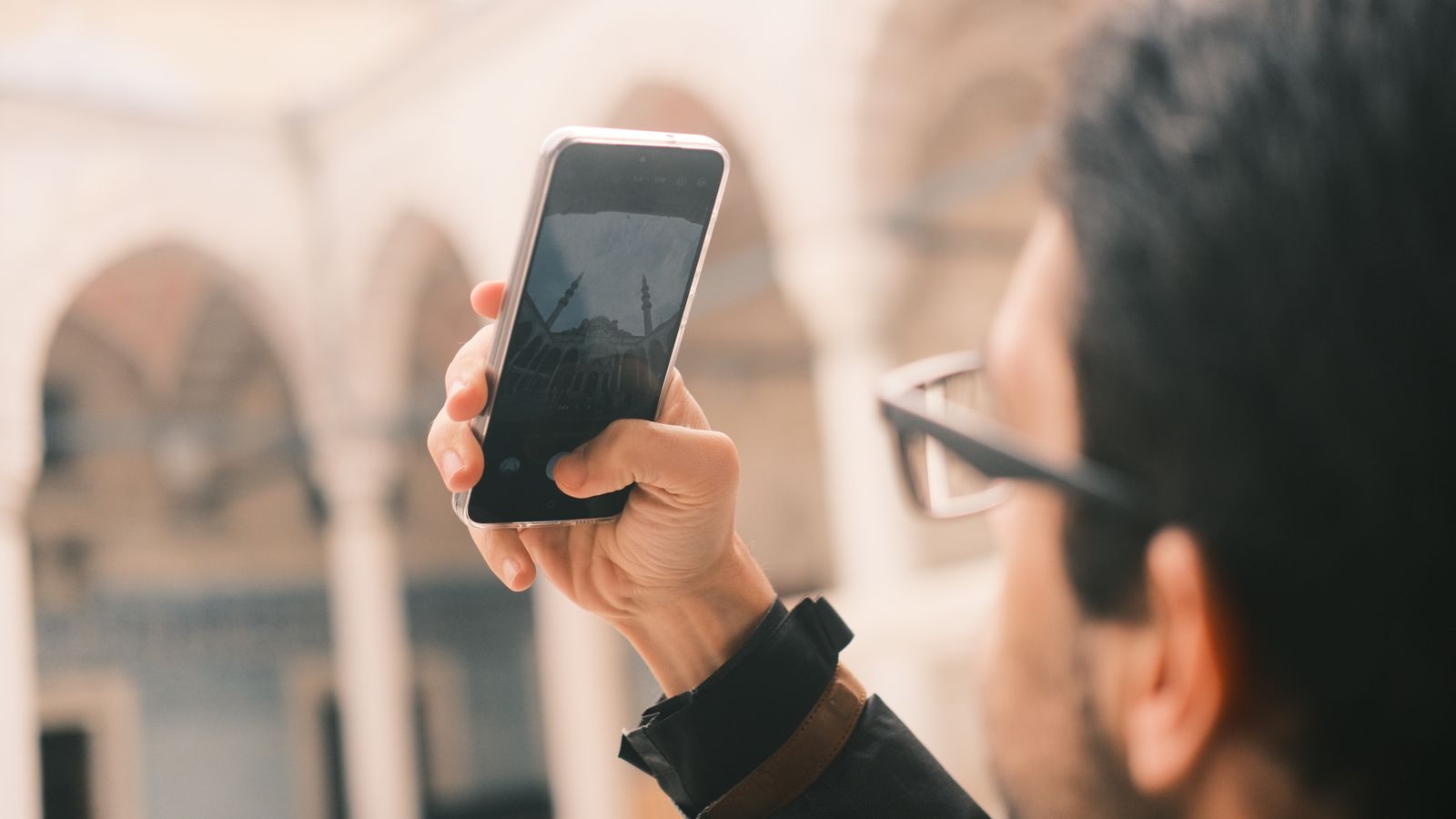 An adult male photographs a mosque using a smartphone outdoors with a focus on capturing architecture.