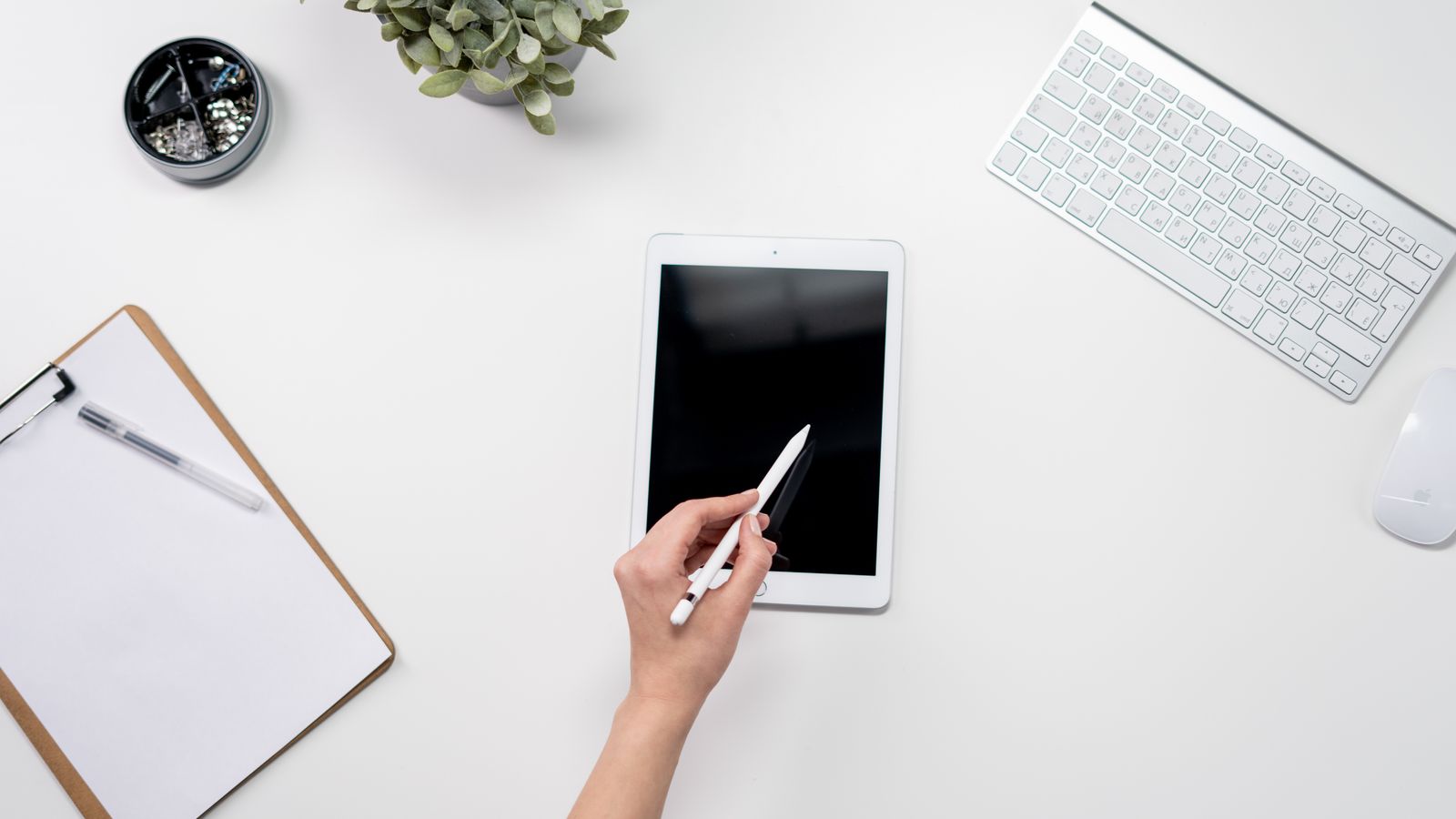 A minimalist home office setup featuring a tablet, stylus, keyboard, plant, and stationery on a white desk.