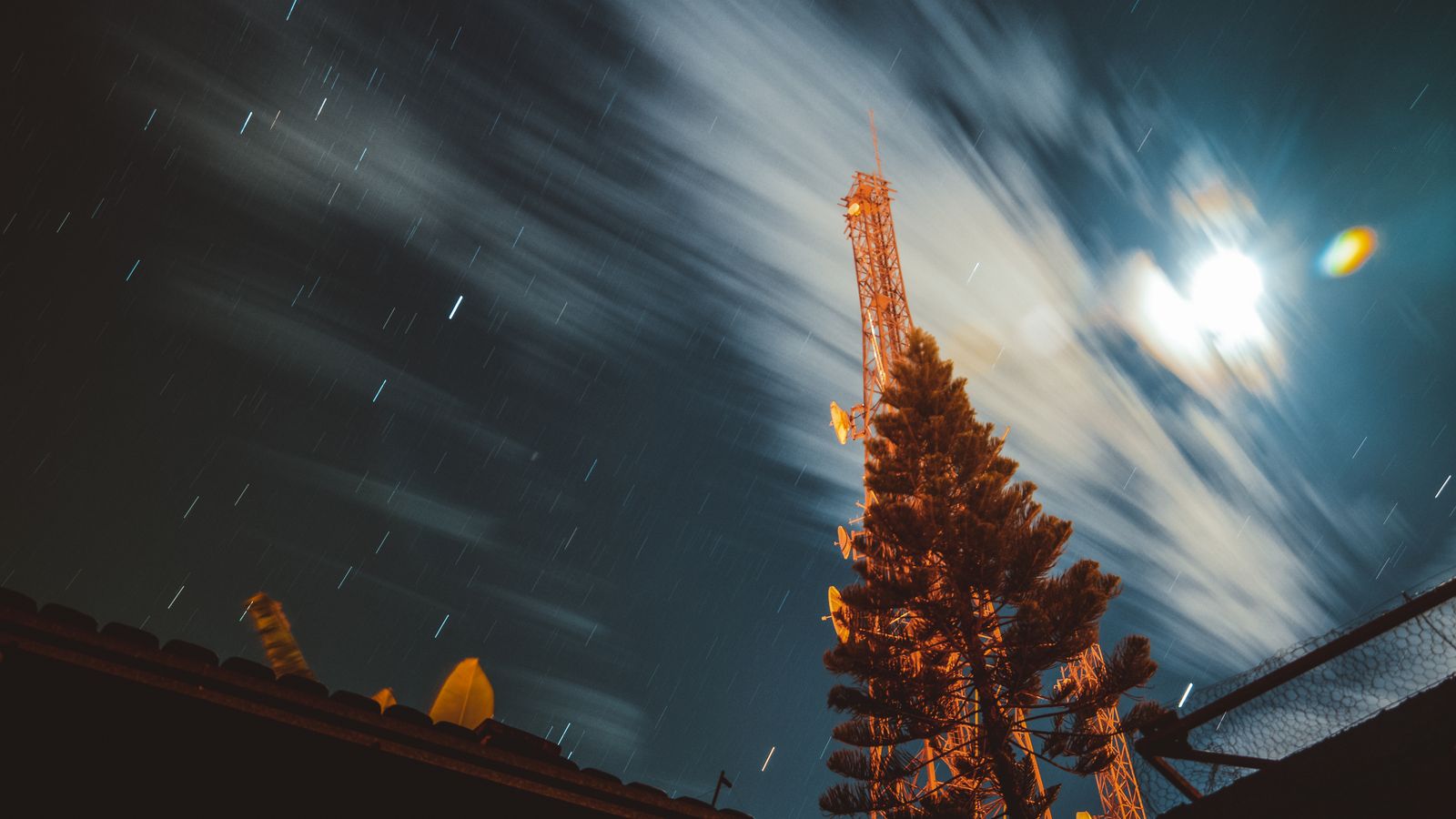 Long exposure of a night sky featuring an antenna with star trails and clouds.