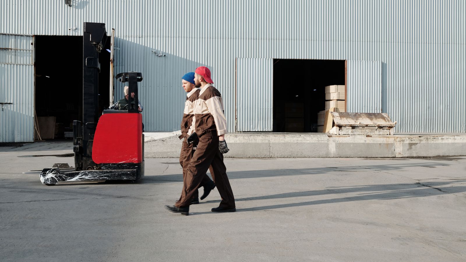 Two workers in uniforms walking outdoors near a forklift at a warehouse.