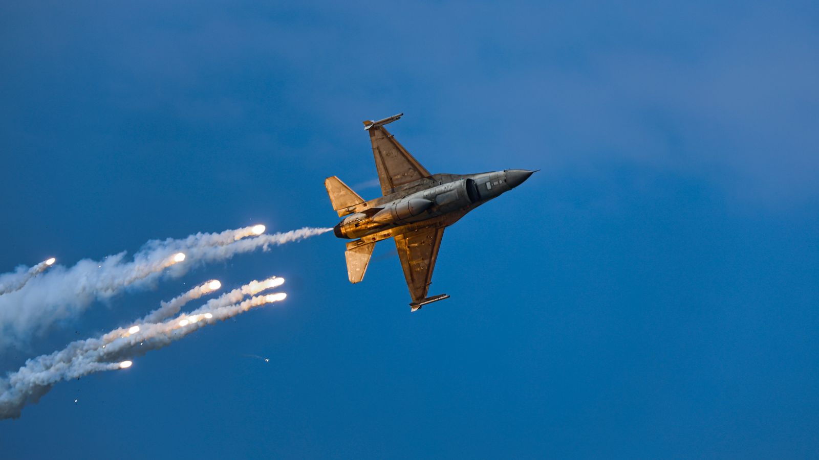 An F-16 fighter jet releases flares while performing an aerobatic maneuver against a clear blue sky at dusk.
