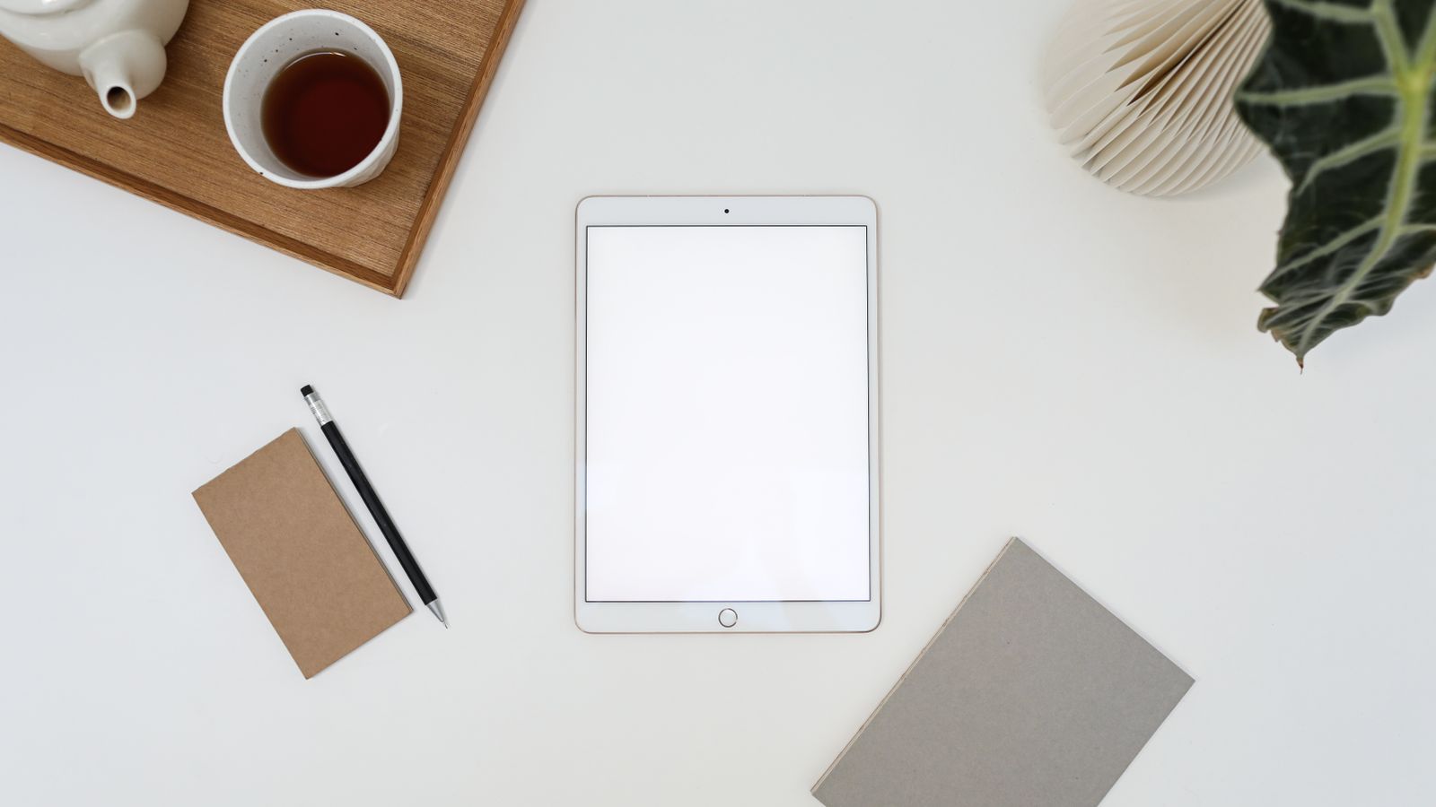 Top view of tablet with empty screen placed near cup of tea and notebook on white table