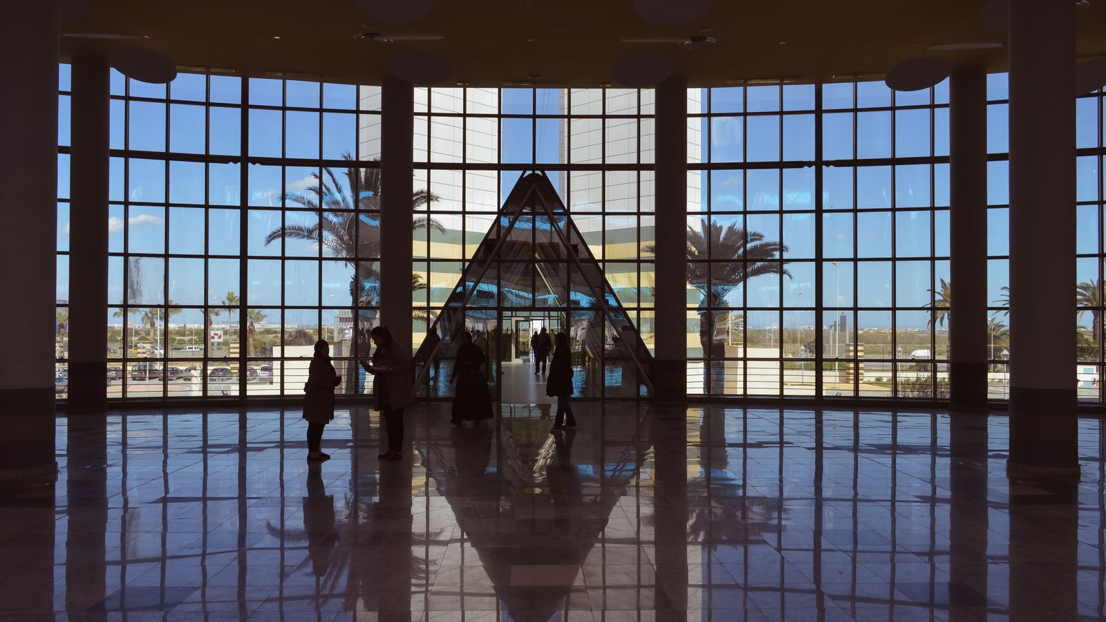 Interior view of a spacious lobby with a central glass pyramid and reflective floor under natural daylight.