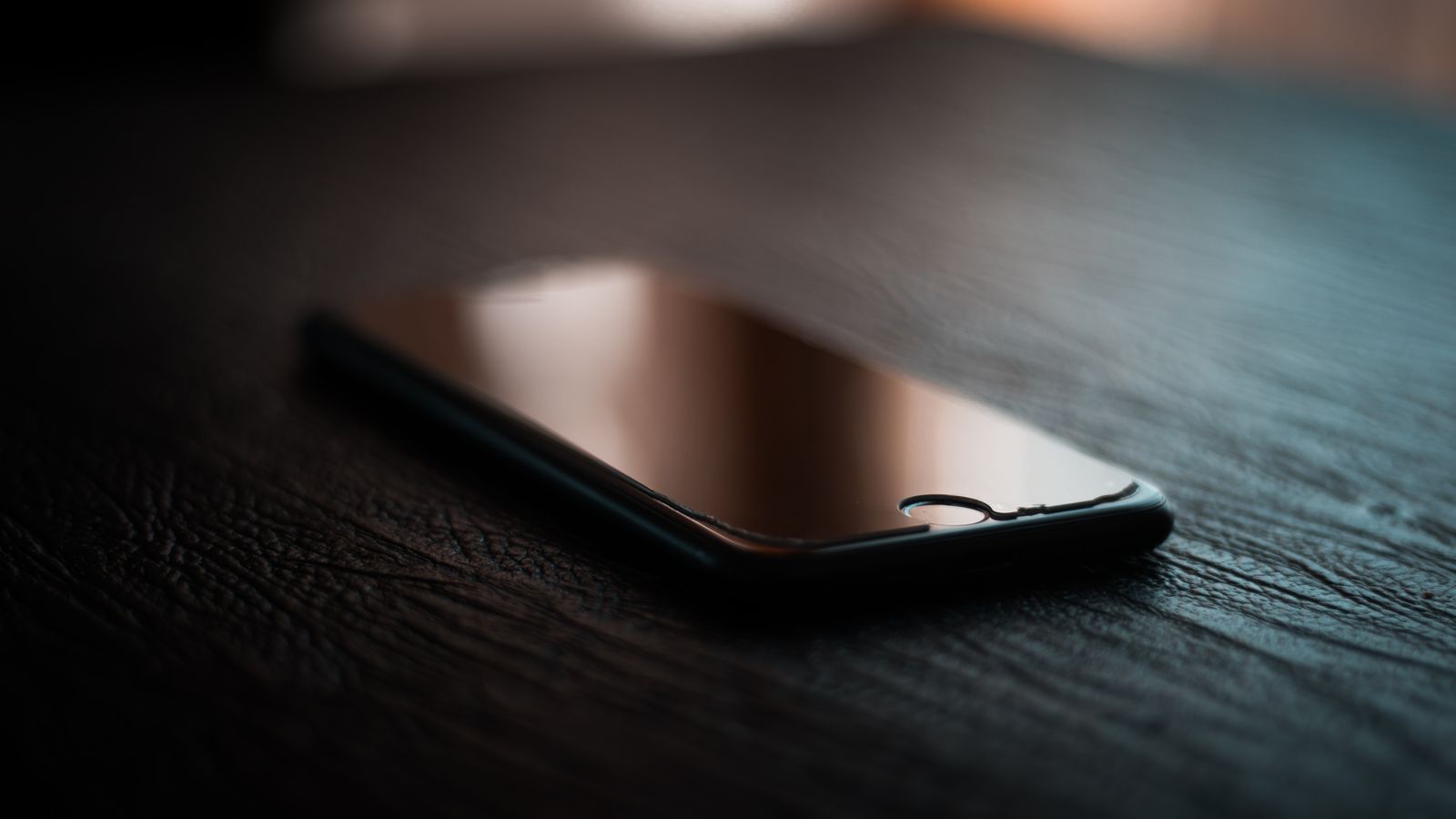 A smartphone lies on a dimly lit wooden table showcasing reflection and shadows.