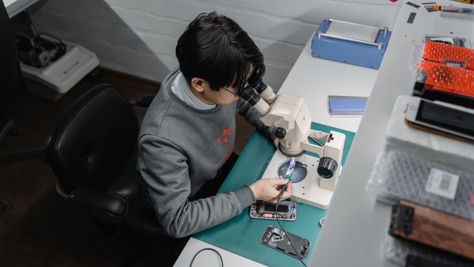 Technician works on smartphone repair under a microscope, indoors at a tech bench.