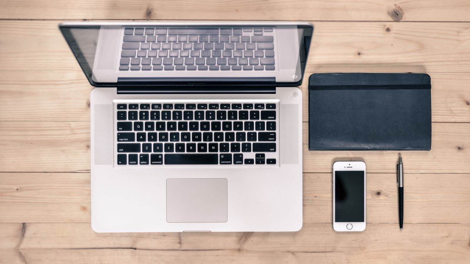 Top view of a tidy workspace featuring a laptop, smartphone, notebook, and pen on a wooden desk.