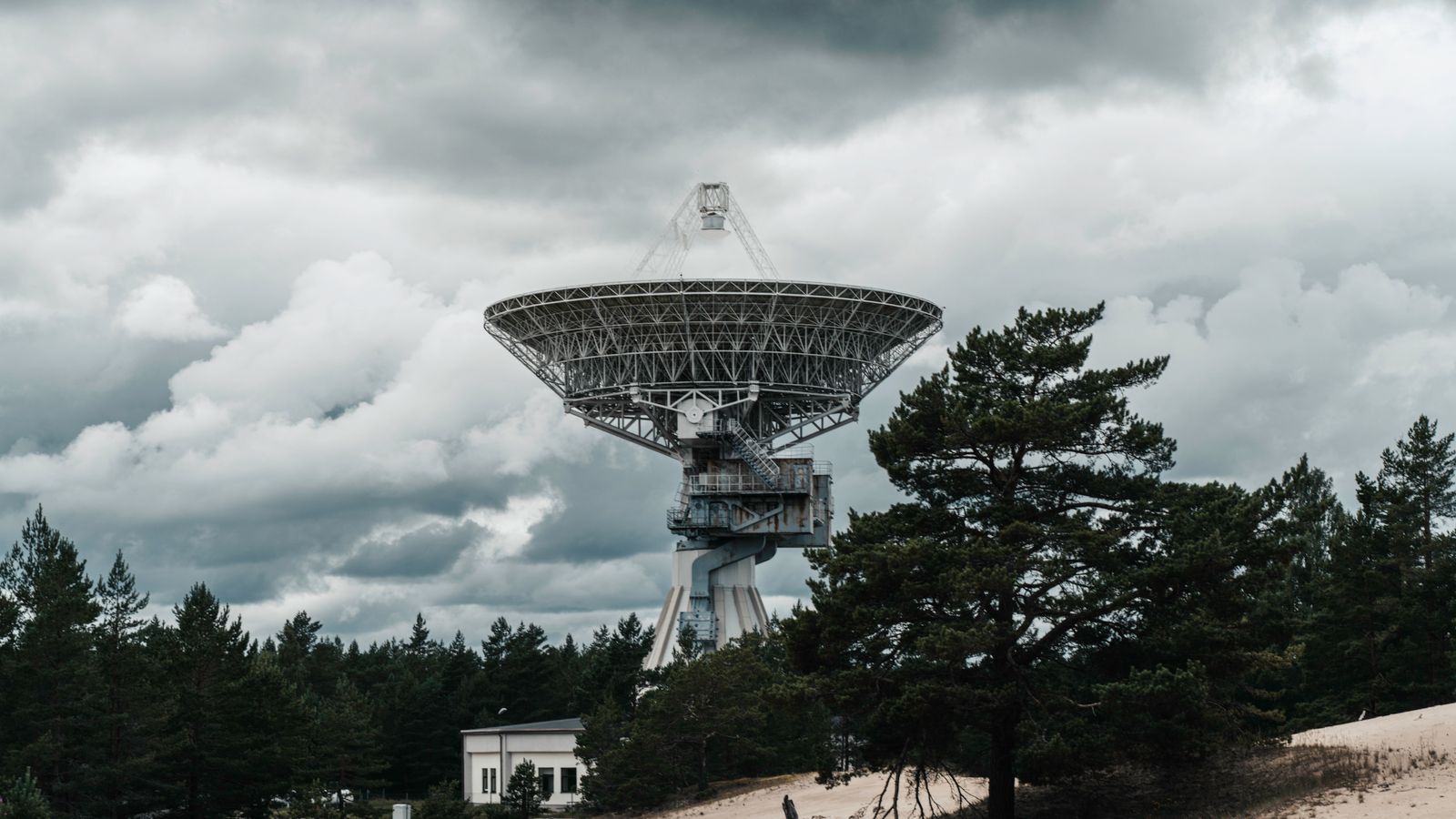 A large radio telescope tower stands in a forest under dramatic clouds, showcasing space technology.