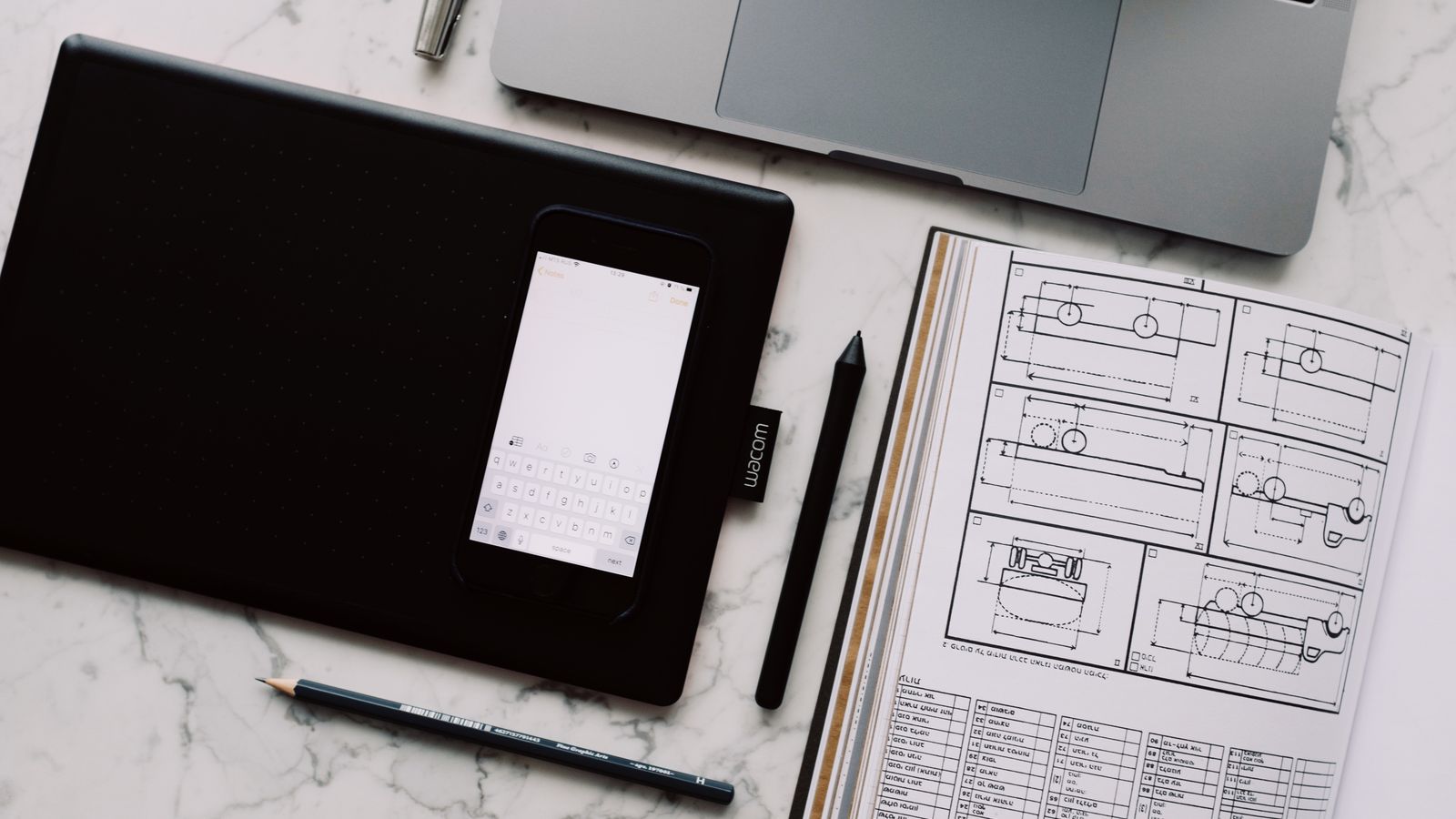 A modern workspace featuring a drawing tablet, smartphone, laptop, and design documents on a marble desk.
